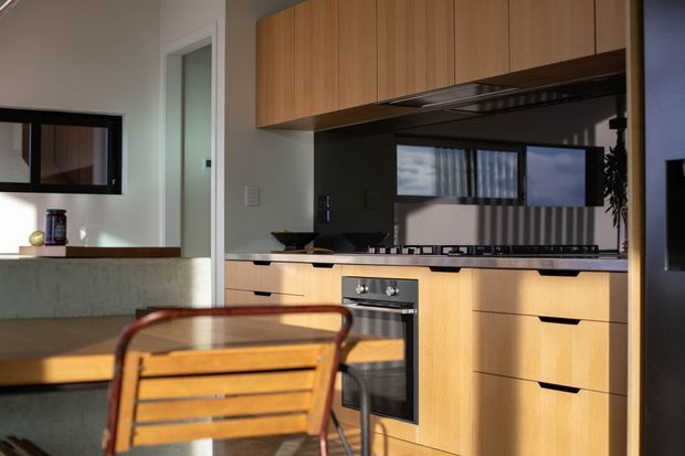 A compact contemporary kitchen with warm timber cabinetry, a dark reflective splashback, pale stone benchtops, integrated oven and cooktop, and a small adjoining dining area. Strong sunlight enters through the windows, creating reflections and shadow patterns across the cabinetry and splashback. A timber dining table and slatted chair occupy the foreground.