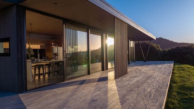 A modern single-level home viewed from a broad timber deck at sunset. Large floor-to-ceiling glazed sliding doors open to a warm interior with a visible kitchen and island seating. Dark vertical timber cladding and slender timber battens frame the glazing, while the deck projects toward a wooded, hilly landscape.