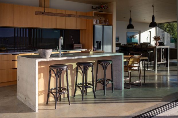A warm, open-plan kitchen with extensive timber cabinetry, a dark reflective backsplash, stainless-steel appliances, and a large concrete island with three bar stools. The island opens toward a dining area with a table, chairs, pendant lights, and large windows. Polished concrete floors and strong late-afternoon sunlight create a relaxed, modern atmosphere.