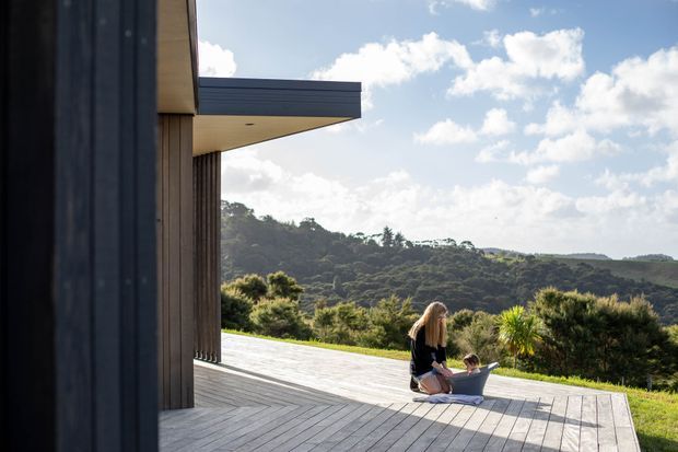Exterior architectural photograph of a modern home opening onto a broad timber deck. Dark vertical cladding and a projecting flat-roofed section frame the left side, while a woman and child sit on the deck overlooking dense rolling bushland. The scene is sunlit and spacious, with a strong indoor-outdoor connection and expansive landscape outlook.