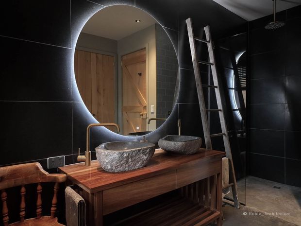 A dramatic, dark-toned bathroom featuring a timber vanity with two vessel basins, brass-toned tapware, and a large circular mirror with integrated backlighting. Black large-format wall tiles continue into the walk-in shower, while a timber ladder and warm wood door add rustic character. A traditional timber balustrade appears in the foreground.