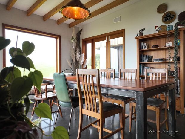 Warm, characterful dining room with a large timber dining table surrounded by mixed timber and upholstered chairs. The room features exposed timber ceiling beams, pale painted walls, timber-framed glazing and doors, dark flooring, and a built-in timber shelving unit displaying books and decorative objects. A copper-toned pendant light provides a prominent focal point, while indoor plants and natural materials add an organic feel.