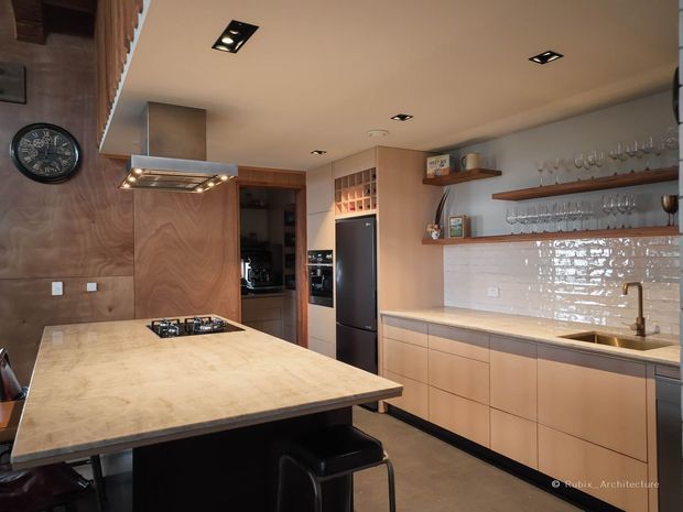 A warm, contemporary kitchen with a large stone island featuring an integrated gas cooktop and seating, flat-panel timber-toned cabinetry, a stainless-steel rangehood, and a full-height bank of appliances. The back wall includes a polished white tiled splashback, long timber display shelves with glassware, and a sink beneath the shelving. Recessed ceiling lighting and timber wall panelling contribute to a refined, functional entertaining space.