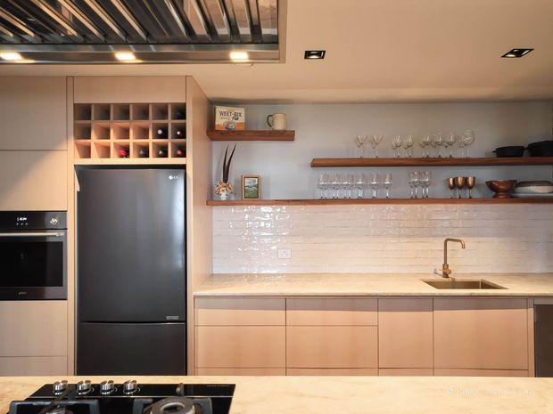 Warm contemporary kitchen viewed from the island, featuring pale timber cabinetry, integrated stainless-steel oven and refrigerator, a white glossy brick-tile splashback, open timber display shelves with glassware, and a stone island benchtop with cooktop. A black-framed glazed ceiling opening or skylight adds an industrial accent.