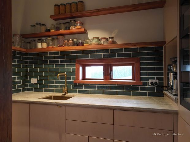 Compact, warm-toned kitchen with flat-panel taupe cabinetry, a pale stone benchtop, brass tapware, dark green subway-tile splashback, and a timber-framed horizontal window. Open timber shelves display jars and kitchen storage, while integrated appliances are visible at the right.
