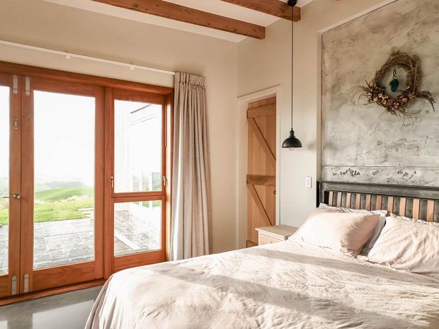A warm, rustic bedroom with a large bed in the foreground, a black metal-and-timber headboard, and a textured decorative wall panel behind it. Full-height timber-framed glazed doors open toward a paved outdoor area and pastoral landscape. Exposed timber ceiling beams, linen curtains, a timber barn-style door, and a hanging black pendant reinforce the relaxed country character.