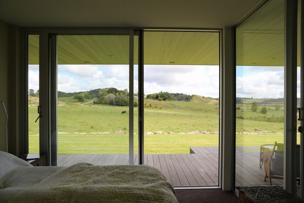 A bedroom photographed from beside the bed, opening through full-height glazed doors onto a timber deck and expansive rolling green countryside. Dark-framed floor-to-ceiling glazing dominates the room, with a bed in the foreground and a small outdoor seating area visible through the side glazing. The interior is simple and subdued, with strong indoor-outdoor connection and rural views.