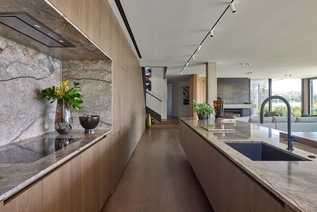 A sophisticated open-plan kitchen and living area viewed along a central circulation path. The kitchen features extensive warm timber cabinetry, a large stone island with an integrated dark sink and sculptural gooseneck tap, and a matching stone-lined display niche with a concealed rangehood. Beyond the kitchen is a bright living space with a pale sofa, floor-to-ceiling glazing, and views of the surrounding urban landscape. A staircase and decorative artwork are visible in the background. The scheme is refined and contemporary with natural stone, timber, and restrained linear lighting.