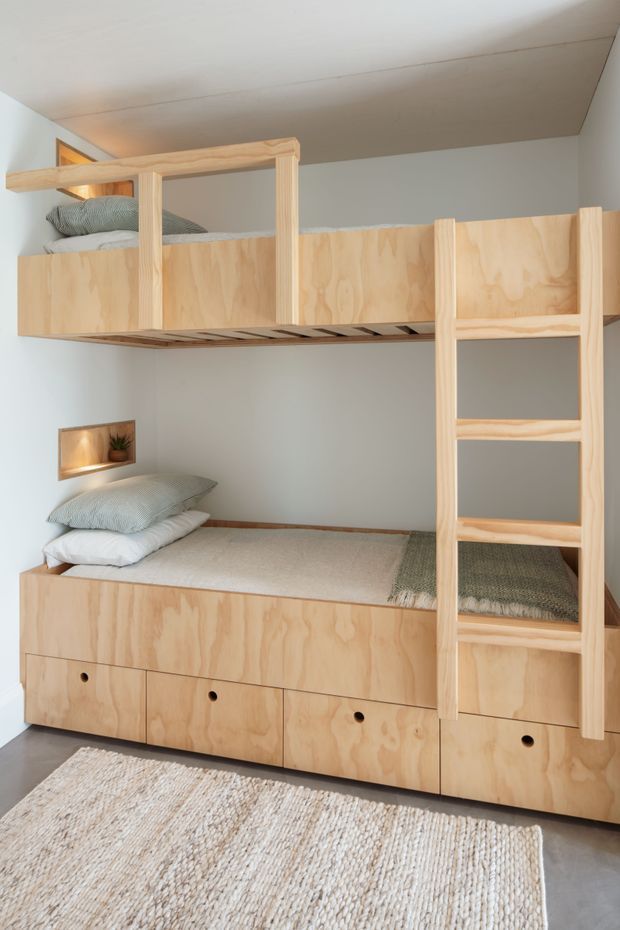 A compact children's bedroom featuring a custom built-in bunk bed and lower bed made from pale plywood. The upper bunk has a timber ladder and guardrail, while the lower bed incorporates generous drawer storage. Neutral bedding, a small recessed wall niche, and a textured woven rug create a simple, warm and functional space.