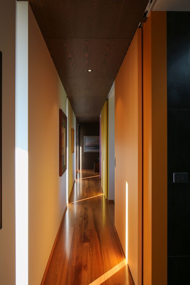 A long, narrow residential hallway with warm orange-painted walls, glossy dark timber flooring, and a dark timber-paneled ceiling with recessed downlights. Framed artwork lines the walls, doorways open along the corridor, and strong sunlight creates bright diagonal bands across the floor and walls. A dark tiled or painted wall is visible at the right edge.