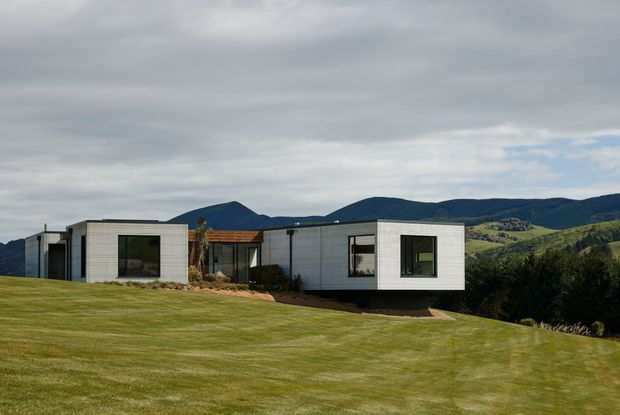 A low-profile modern single-storey house set within a broad manicured lawn, with rectilinear white-clad volumes, dark-framed windows, and a small timber-lined entry or connecting section. Rolling forested hills and mountains form the background beneath an overcast sky.