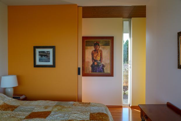A warmly lit bedroom with a low bed in the foreground, an ochre-orange feature wall, framed artwork, timber ceiling paneling over the doorway, polished timber flooring, and a narrow vertical window providing a view outside. A dark timber dresser occupies the lower right corner.