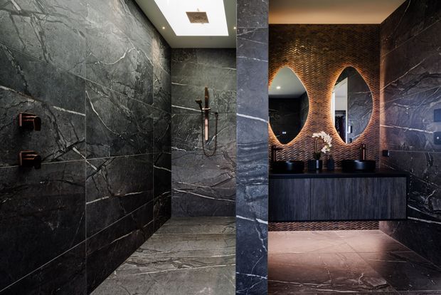 A dramatic, high-end bathroom clad in dark veined stone, with a walk-in shower on the left and a floating dark timber vanity on the right. Sculptural arched mirrors and a textured bronze-toned feature wall create a bold, moody interior.