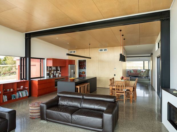 Open-plan contemporary living space with a dark leather sofa in the foreground, dining area and compact kitchen beyond. The room features exposed black steel framing, warm timber wall and ceiling panels, red built-in cabinetry, polished concrete or terrazzo-style flooring, large windows, and a fireplace wall at right. A further seating area is visible through the opening at the rear.