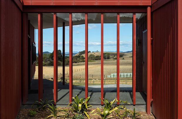 View from within a red-toned architectural structure toward a rural agricultural landscape. Tall floor-to-ceiling glazed openings are divided by slender red vertical mullions, with dark red ribbed wall cladding and a matching overhead structure. A narrow planted bed occupies the foreground, while a paved terrace and expansive fenced fields are visible beyond.