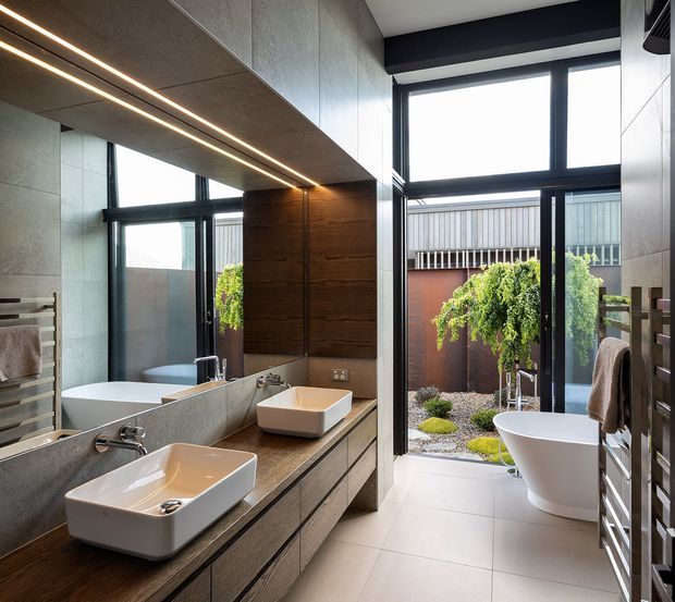 Contemporary bathroom with a double vanity, large frameless mirror, freestanding bathtub, full-height black-framed glazing, and a private landscaped courtyard visible beyond. The room features a restrained palette of grey stone-look surfaces, dark timber cabinetry, black metal frames, and warm integrated linear lighting.