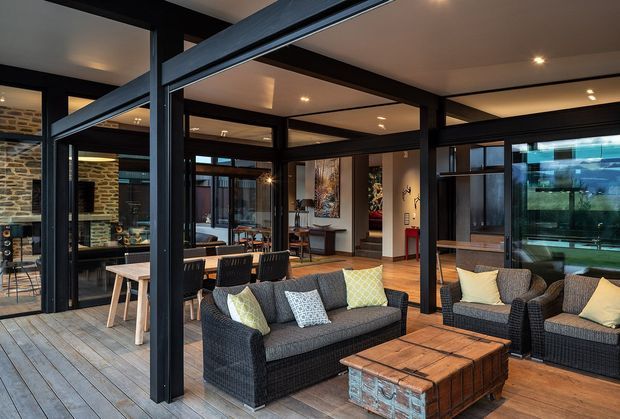 A contemporary indoor-outdoor living area with dark exposed steel or metal-framed glazing and structural posts, timber flooring, and extensive glass partitions. Wicker-style sofas and armchairs surround a rustic timber coffee table, while a dining area and kitchen are visible beyond. The space has warm recessed lighting and a strong connection to the exterior through large glazed openings.