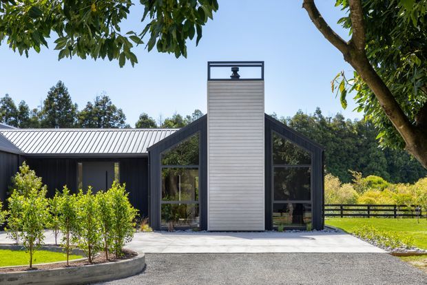 Exterior view of a contemporary single-storey rural-style home with a dark charcoal gabled facade, large glazed openings, and a central light horizontal-clad chimney or entry tower. The building has a silver-grey pitched metal roof, generous glazing, and a broad concrete forecourt, set within landscaped grounds with hedges, mature trees, and fencing.