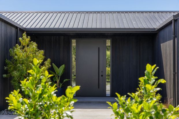 A modern residential entry facade viewed from the front walkway. The entrance is framed by dark vertical cladding and a projecting corrugated metal gable roof, with a tall charcoal door and narrow glazed sidelights. Lush foreground planting partially screens the concrete path and threshold.