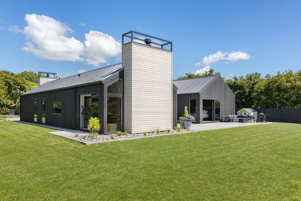 Exterior view of a contemporary single-storey home set in a landscaped lawn. The building combines dark vertical cladding with a lighter horizontal-clad central tower, large glazed openings, and simple pitched metal roofs. A paved terrace with outdoor furniture is visible beside the right-hand wing, with trees and a fenced boundary behind.