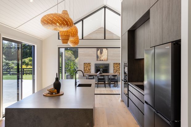 A bright open-plan kitchen viewed from the island, with a large grey stone island, dark integrated cabinetry, stainless-steel refrigerator, light timber flooring, and a white vertical-panel ceiling. Black-framed glazing opens toward a landscaped exterior, while the rear dining area includes a fireplace wall with timber detailing. Sculptural woven pendant lights hang above the island.