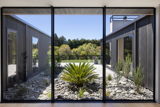 View from inside through large black-framed glazed doors toward a landscaped courtyard between two dark vertical-clad wings. The courtyard features pale river stones, a central architectural plant, sparse grasses, and a low dark fence, with a bright open sky beyond. The interior has a white slatted ceiling and pale flooring visible at the edges. The composition emphasizes strong indoor–outdoor connection and a contemporary courtyard setting.