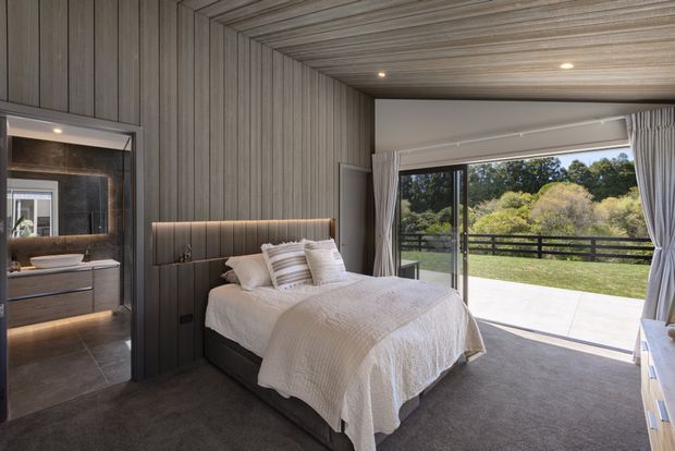 A refined contemporary bedroom with a centered upholstered bed, layered neutral bedding, dark vertical wall panelling, and a matching timber-look slatted ceiling. A softly illuminated integrated headboard provides accent lighting. Full-height glazed doors open to a paved terrace and landscaped garden, while a bathroom or dressing area is visible through the opening on the left.