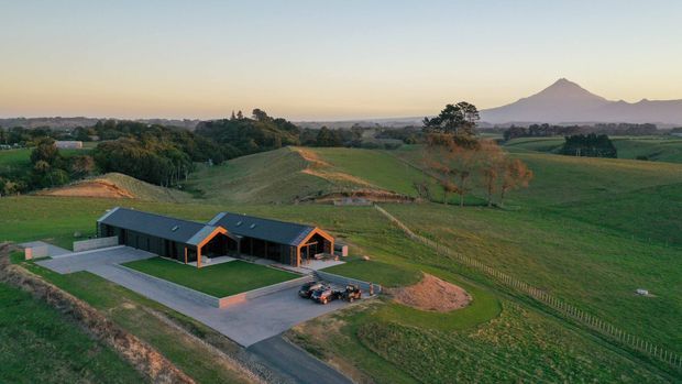Aerial view of a contemporary low-profile rural property set amid expansive green farmland, with dark cladding, orange-framed gabled forms, paved outdoor areas, and a prominent conical mountain in the distance.