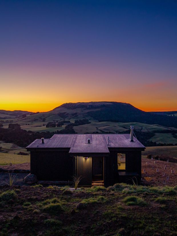 A small dark-clad cabin set in an open rural, mountainous landscape at dusk. The compact single-storey building has a corrugated metal pitched roof, simple rectangular form, a central recessed entry, small windows, and a visible chimney. Warm interior light contrasts with the purple-blue sunset and rolling farmland beyond.