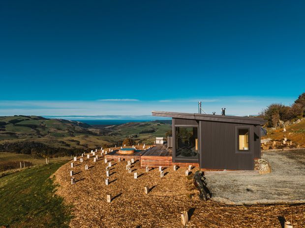 A compact dark-clad cabin or small dwelling sits on a rugged elevated rural site, with a flat or very low-pitched roof, timber deck, large windows, and a gravel approach. The foreground includes a landscaped gravel and timber-post area, while rolling green hills and a distant blue ocean or coastal horizon are visible beyond. The scene is photographed in clear daylight with a strong blue sky.