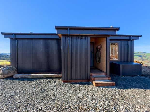 A compact modern single-storey building set on a rocky, elevated rural landscape. The exterior is clad in dark vertically ribbed metal, with a low-pitched flat roof, simple rectilinear forms, and a recessed timber-lined entry with steps. A narrow window and projecting side volume add articulation to the facade.