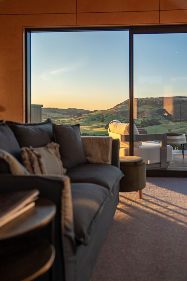 A warm, contemporary living room with a large dark upholstered sectional sofa, soft cushions, a round side table, and carpeted flooring. Floor-to-ceiling glazed doors or windows open to an expansive rural valley and hills, with sunset light casting long shadows across the room. Timber-toned wall panelling frames the glazing, and an outdoor seating area is visible beyond.