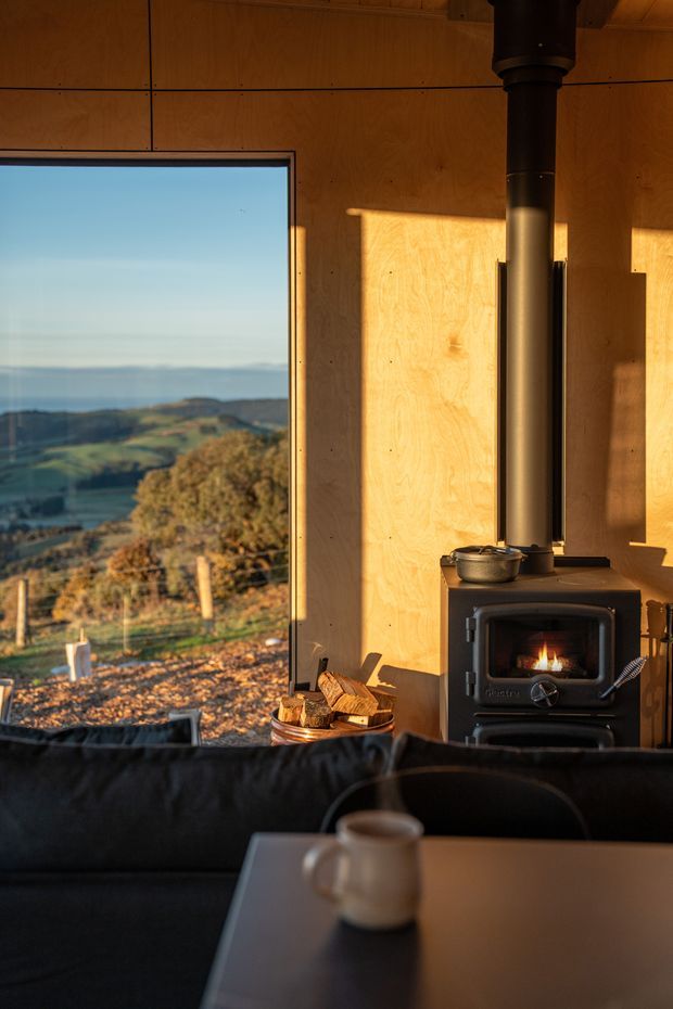 Warm, atmospheric interior of a compact timber-lined living room or cabin, featuring a black wood-burning stove with visible fire, a dark sofa, and a table in the foreground. A large window frames a scenic coastal or rural landscape, while low golden sunlight creates strong warm highlights and shadows across the plywood walls.