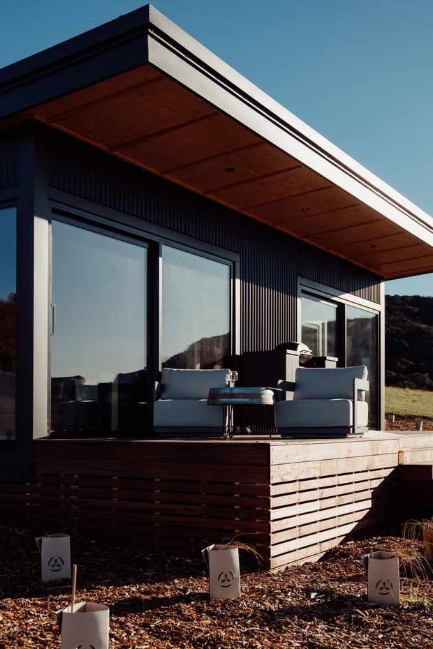 Exterior view of a contemporary single-storey dwelling with a projecting flat roof, dark vertical ribbed cladding, large sliding glass doors, and a timber-lined soffit. A raised timber deck features outdoor lounge seating and a small table, with a landscaped mulched foreground and natural hillside setting.