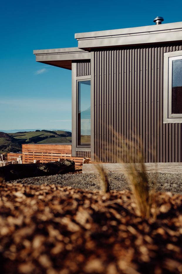 Exterior close-up of a compact modern building with a flat roof, strongly ribbed dark vertical cladding, light-coloured window and door frames, and a timber deck or railing. The structure sits in a rural, elevated landscape with rolling farmland visible beyond. Bright clear daylight and low-angle photography emphasize the textured facade and roof forms.