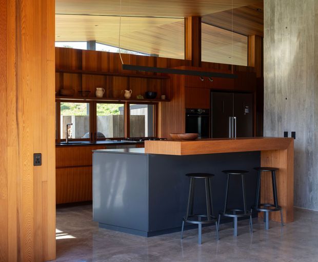 A warm, contemporary kitchen with extensive vertical timber wall and ceiling lining, dark integrated cabinetry, two substantial kitchen islands, and black bar stools. A large black linear pendant hangs above the island, while a polished concrete wall and concrete-look floor add contrast. Timber-framed windows provide natural light and views to the garden or courtyard.