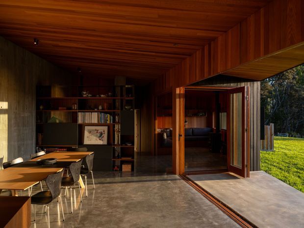 Warm, dark timber-lined open-plan interior with a dining area in the foreground and a connected living space beyond. Built-in shelving and cabinetry line the rear wall, while large timber-framed folding or sliding doors open directly onto a lawn and landscaped exterior. A polished concrete floor and timber ceiling create a strong indoor-outdoor connection.