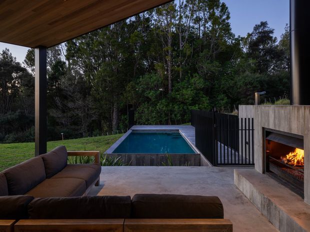Covered outdoor entertaining area with a dark sectional sofa, concrete paving and a timber-lined ceiling, overlooking a compact swimming pool and dense garden. A built-in concrete outdoor fireplace with a lit fire sits to the right, beside black vertical metal fencing and a timber-toned privacy wall.