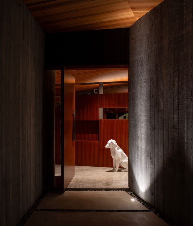 A dramatic, dimly lit architectural interior viewed down a narrow passage toward a richly timber-lined room. Dark textured wall panels frame the corridor, while warm timber cladding covers the ceiling and rear walls. Slab-like floor paving leads into the space, where a faceted white animal sculpture provides a bright focal point. Integrated lighting creates strong contrasts and a refined, gallery-like atmosphere.