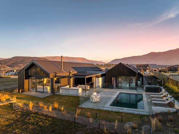 A contemporary single-storey residence set in a dry, mountainous landscape at sunset. The home comprises several connected gabled volumes with dark timber or metal-clad walls, extensive floor-to-ceiling glazing, pitched metal roofs, and warm interior lighting. A paved terrace, rectangular swimming pool, outdoor seating, and landscaped native grasses surround the house.