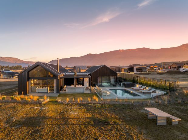 Wide exterior view of a contemporary single-storey house in a mountainous semi-rural setting at dusk. The home has dark vertical cladding, steep pitched gabled roof forms, extensive floor-to-ceiling glazing, a terrace and rectangular swimming pool, with warm interior lighting and landscaped planting around the building.