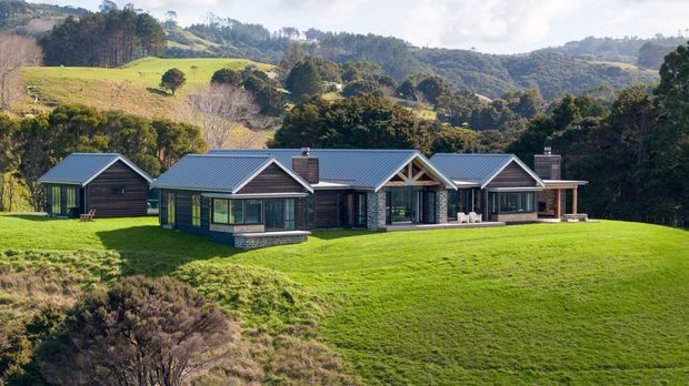 A low, sprawling single-storey rural home set in a broad manicured lawn, with multiple connected gabled wings, dark timber-clad walls, blue-grey metal pitched roofs, large windows, and stone base detailing. Rolling green hills and wooded countryside form the background.