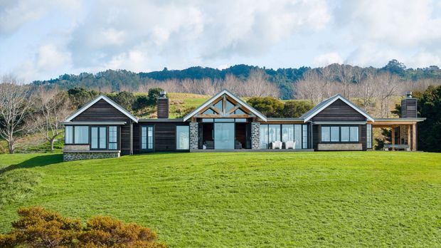 Single-storey symmetrical contemporary residence set in a broad landscaped lawn, with three connected gabled sections, dark horizontal cladding, extensive glazing, stone accents, and covered outdoor living areas. Rolling rural greenery and wooded hills form the background.