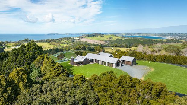 Aerial view of a sprawling single-storey contemporary residence set within expansive manicured lawns and dense mature vegetation. The home has multiple low-pitched gabled roof volumes, dark vertical cladding with light metal roofing, and broad glazed openings. A curving driveway and landscaped grounds lead toward the house, with a sweeping coastal and estuarine landscape visible beyond.