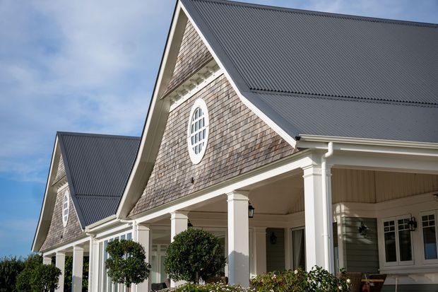 Exterior view of a large traditional residential building with prominent steep gables, dark corrugated metal roofing, shingle-clad gable faces, white-painted columns and trim, and covered verandah or porch areas. Oval and rectangular multi-pane windows and neatly clipped shrubs reinforce the formal, coastal-inspired character.