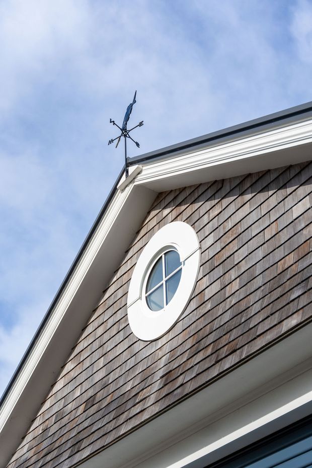 Close-up upward view of a traditional gabled building facade with weathered cedar shingle cladding, crisp white decorative trim and eaves, and a distinctive round window with white surround and cross muntins. A small weather vane is mounted at the roof ridge against a pale blue sky.