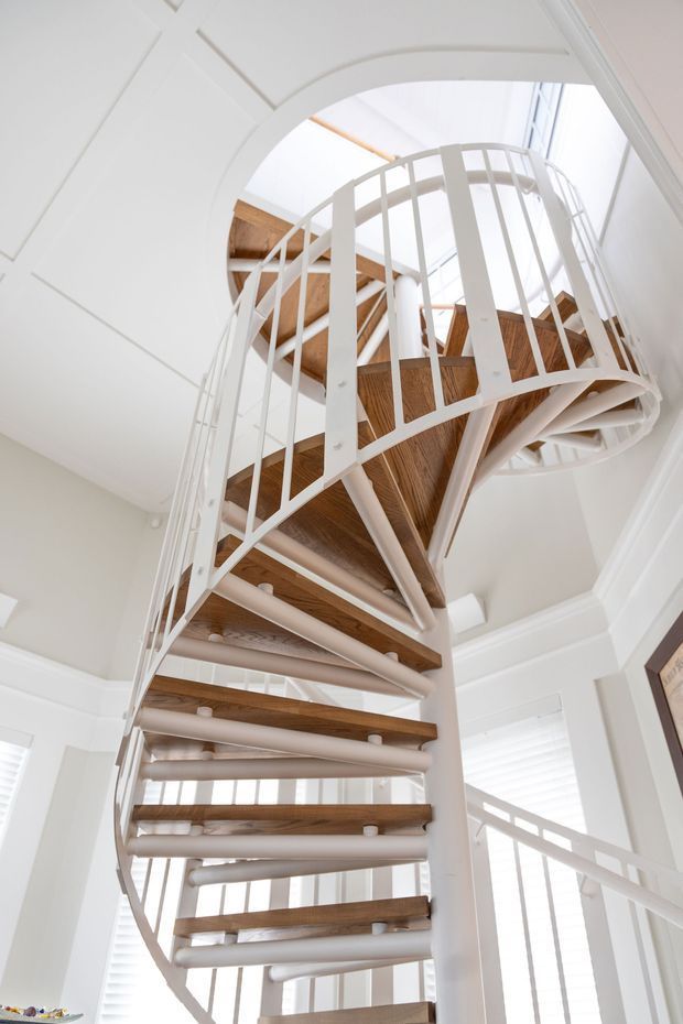 Bright interior view looking up through a sculptural spiral staircase with white-painted metal balustrades and supports, warm timber treads, and a tall open stairwell. White walls, decorative ceiling mouldings, and abundant daylight create an airy, refined setting.