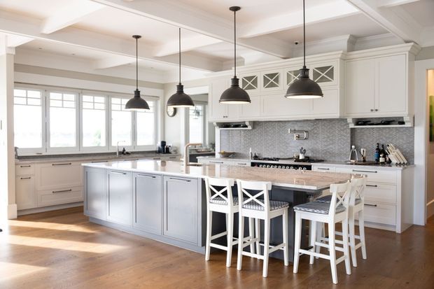 Bright, spacious white kitchen with a large central island, integrated seating, shaker-style cabinetry, patterned tile splashback, timber flooring, and multiple black pendant lights. The room features a coffered ceiling, extensive windows, and a refined classic coastal-inspired fit-out.