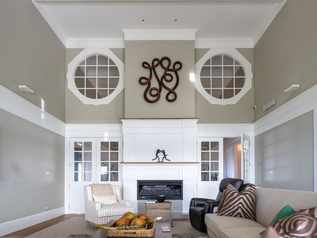 A bright, refined living room with a tall coffered ceiling, white traditional-style fireplace and mantel, built-in glazed cabinetry, circular multi-pane windows, and pale grey walls. Layered seating, patterned cushions, and a central coffee table create a comfortable yet elegant interior.