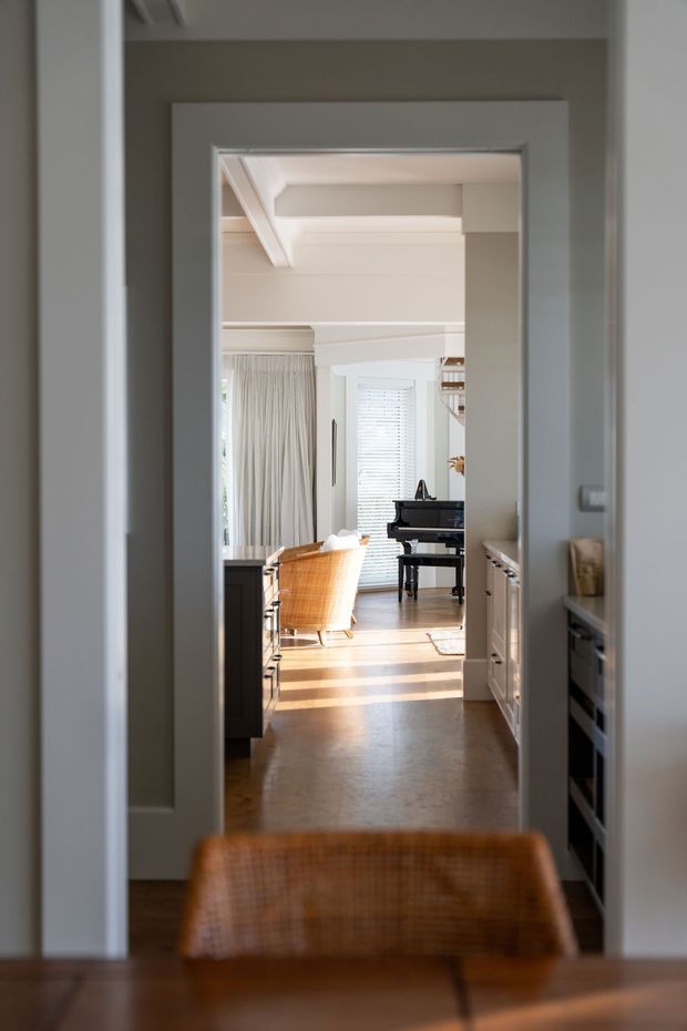 A view through a framed interior doorway and hallway into a bright living area. The space features warm timber flooring, white cabinetry, a black upright piano, woven orange-brown lounge seating, plantation shutters, sheer curtains, and a stepped tray-style ceiling with exposed white beams. The foreground includes part of a timber dining table and woven chair.
